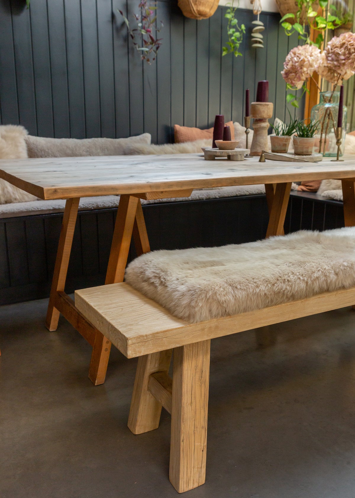 A light natural wood dining table with A-frame legs, paired with a matching bench and a long upholstered bench seat pad in cream sheepskin, set in a rustic dining room with dark gray paneled walls.