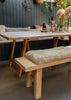 A light natural wood dining table with A-frame legs, paired with a matching bench and a long upholstered bench seat pad in cream sheepskin, set in a rustic dining room with dark gray paneled walls.