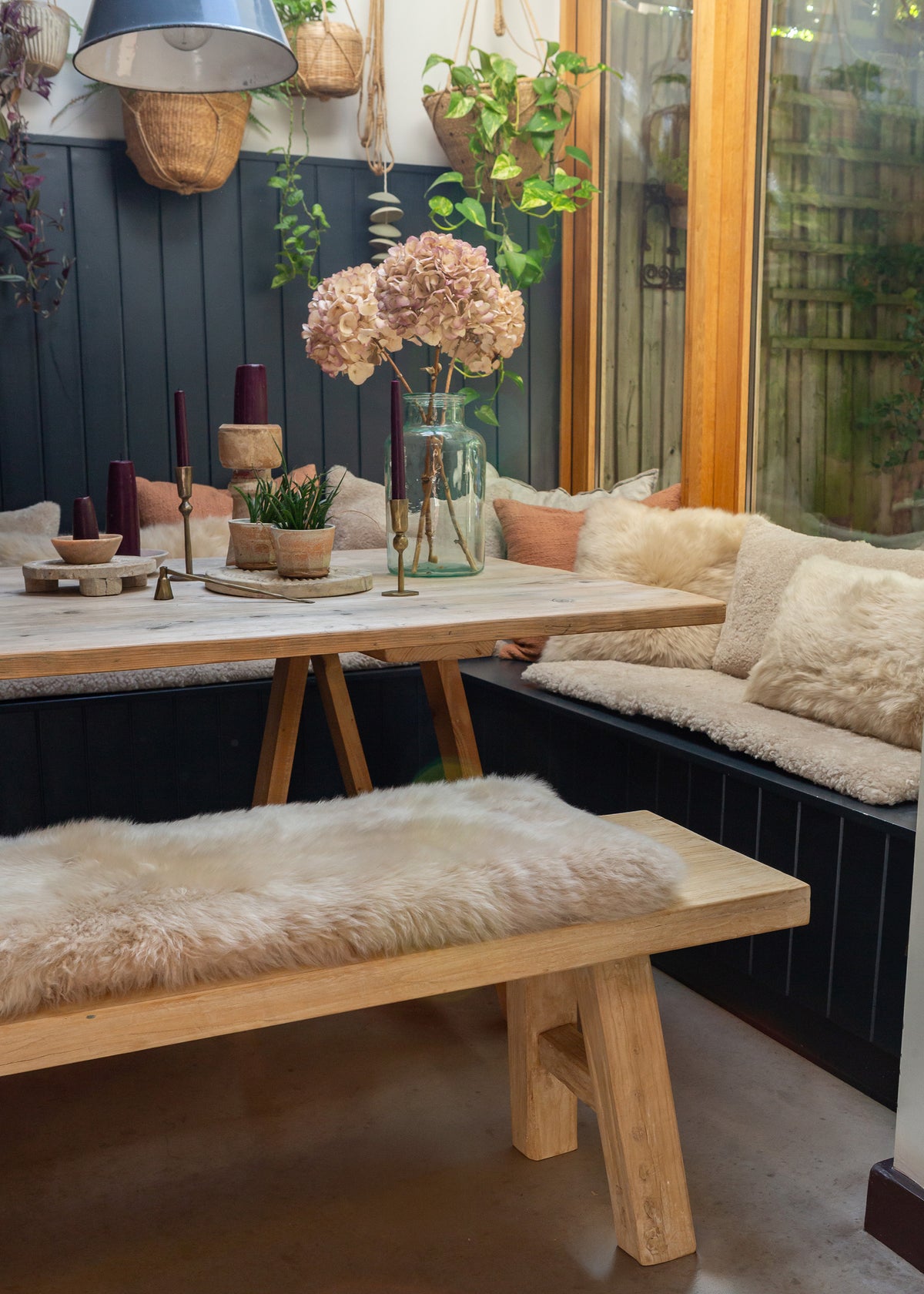 A rustic wooden dining set featuring a table and matching bench with fluffy cream sheepskin cushions, styled in a cozy indoor garden setting with plants and a window.