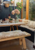 A rustic wooden dining set featuring a table and matching bench with fluffy cream sheepskin cushions, styled in a cozy indoor garden setting with plants and a window.