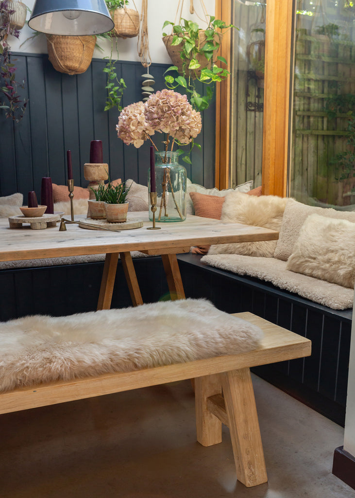 A rustic wooden dining set featuring a table and matching bench with fluffy cream sheepskin cushions, styled in a cozy indoor garden setting with plants and a window.