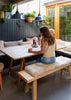 A woman sits at a rustic wooden dining table with a long bench pad in fluffy sheepskin, set in a cozy indoor-outdoor dining space with plants and hanging pendant lights.