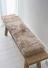A fluffy beige sheepskin bench pad laid across a light wood bench in a bright, minimalist room with white walls and floorboards.