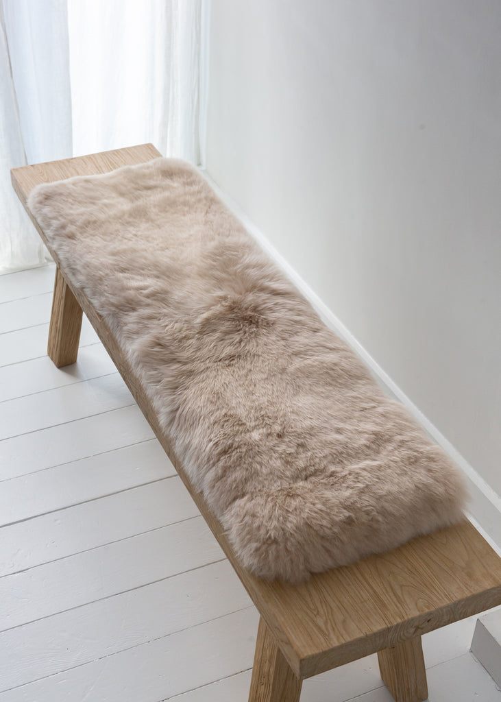A fluffy beige sheepskin bench pad laid across a light wood bench in a bright, minimalist room with white walls and floorboards.