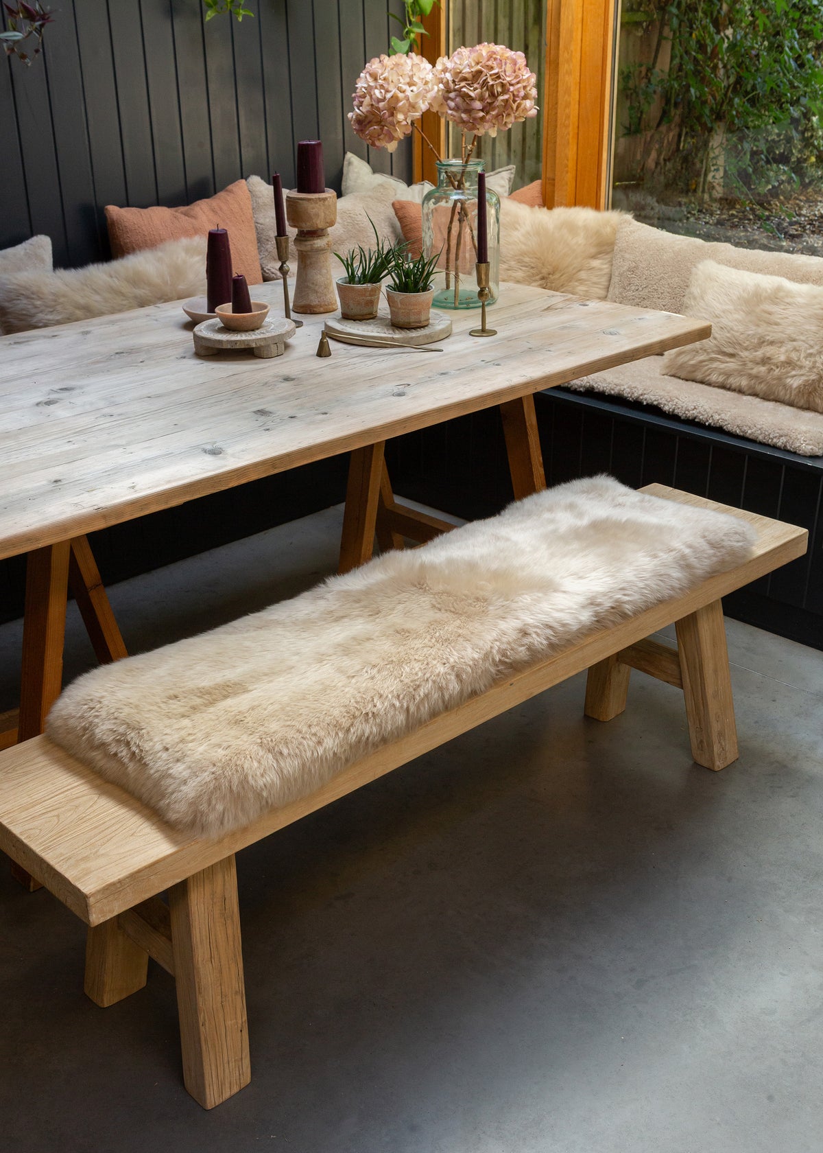 A rustic wooden dining table with a matching bench, with beige sheepskin bench pad, set in an outdoor patio dining area with a built-in bench and cushions behind.