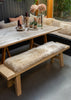 A rustic wooden dining table with a matching bench, with beige sheepskin bench pad, set in an outdoor patio dining area with a built-in bench and cushions behind.