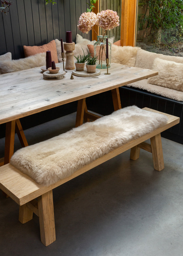 A rustic wooden dining table with a matching bench, with beige sheepskin bench pad, set in an outdoor patio dining area with a built-in bench and cushions behind.