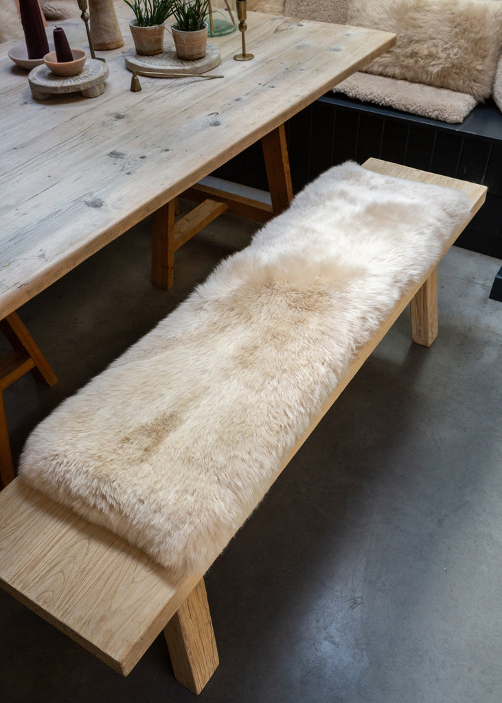 A light natural wood bench with a fluffy cream sheepskin seat cushion, shown in a rustic dining room setting next to a matching wooden table.