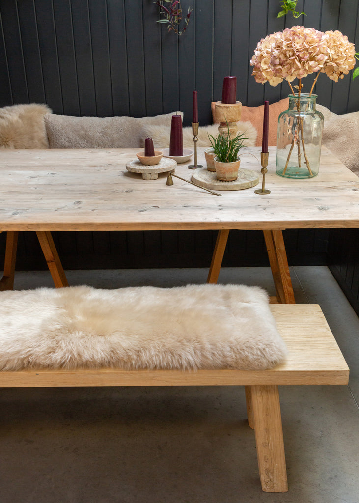 A rustic wooden dining table with matching bench seating, styled with a beige sheepskin bench pad, candles, small potted plants, and a large vase of pale pink hydrangeas against a dark paneled wall.
