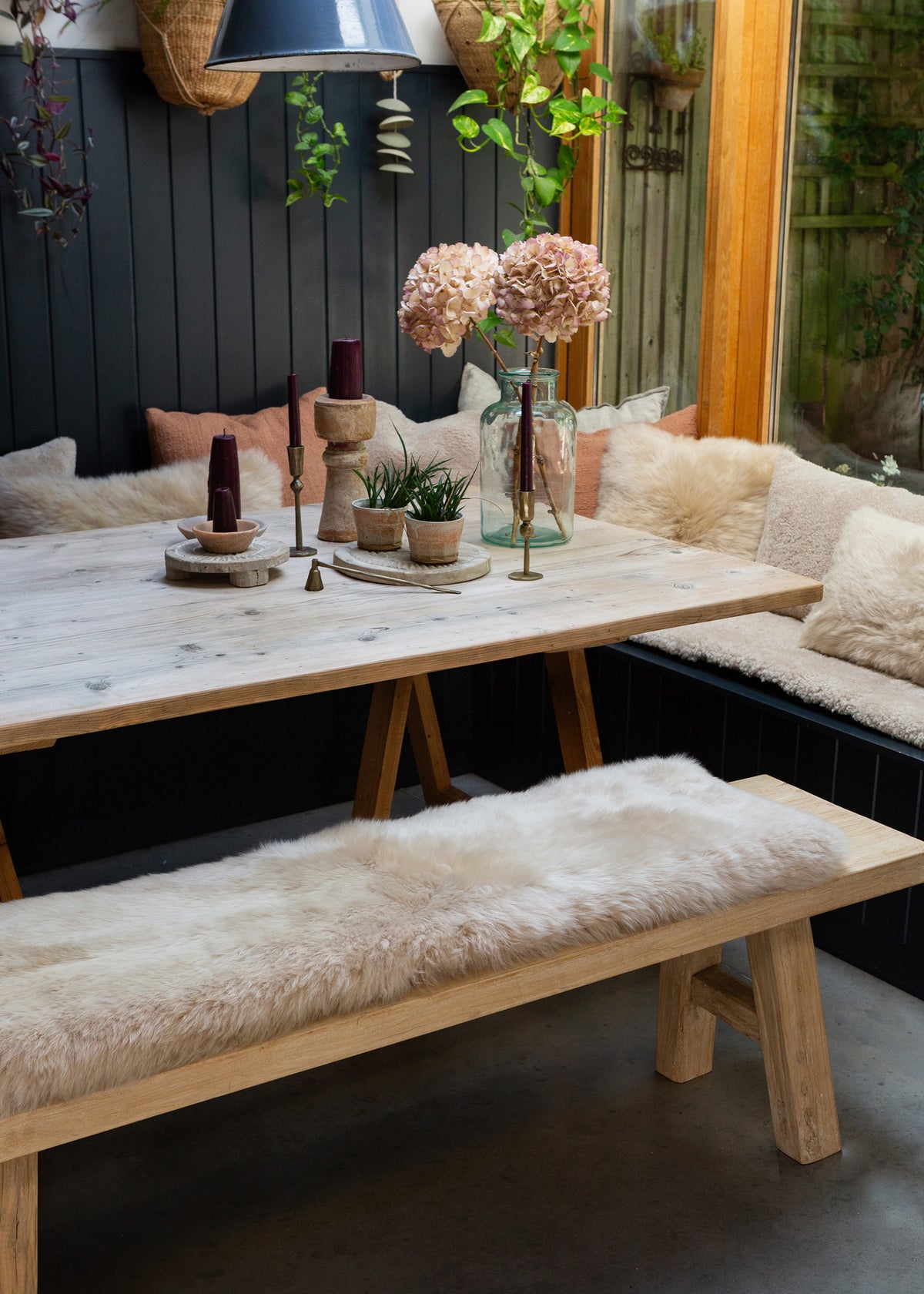 A rustic wooden dining table with A-frame legs, paired with a matching bench and a sheepskin bench pad, set in a cozy outdoor dining area with a dark paneled wall and greenery.