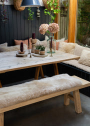 A rustic wooden dining table with A-frame legs, paired with a matching bench and a sheepskin bench pad, set in a cozy outdoor dining area with a dark paneled wall and greenery.