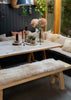 A rustic wooden dining table with A-frame legs, paired with a matching bench and a sheepskin bench pad, set in a cozy outdoor dining area with a dark paneled wall and greenery.