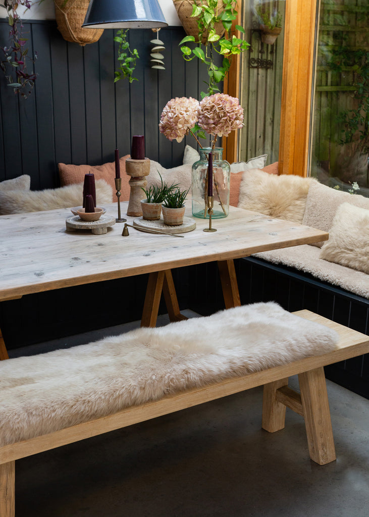 A rustic wooden dining table with A-frame legs, paired with a matching bench and a sheepskin bench pad, set in a cozy outdoor dining area with a dark paneled wall and greenery.