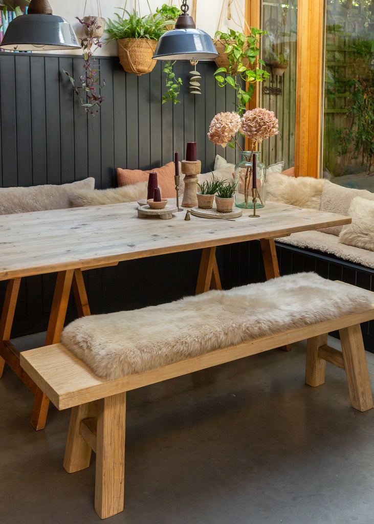 A rustic wooden dining table with matching bench with a sheepskin bench pad, styled in a modern rustic dining room setting with hanging pendant lights and plants.