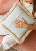 A square throw pillow in a pale sage green linen fabric, framed with a fluffy cream sheepskin trim, held by a woman sitting on a dusty pink velvet sofa.