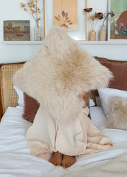 A person sitting on a bed wrapped in a soft cream blanket, holding a large fluffy beige sheepskin pillow in front of their face. The bedroom has white bedding, a woven headboard, and wall art above.