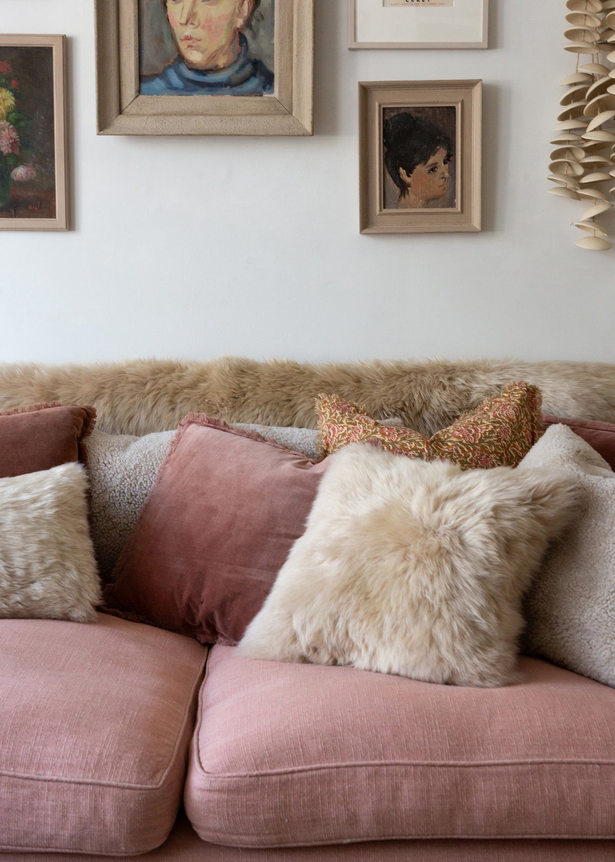 A dusty pink sofa in a styled room setting, decorated with a variety of textured throw pillows including sheepskin and velvet in pink, beige, and rust tones, with framed art on a white wall behind it.