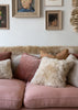 A dusty pink sofa in a styled room setting, decorated with a variety of textured throw pillows including sheepskin and velvet in pink, beige, and rust tones, with framed art on a white wall behind it.