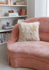 A square cream sheepskin pillow resting on a dusty pink velvet armchair in a bright, modern room with white bookshelves in the background.