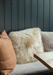 A fluffy cream sheepskin throw pillow centered on a textured beige sofa, flanked by a tan textured pillow on the left and a neutral pillow on the right, set against a dark gray vertical wood-paneled wall with a wooden table edge in the foreground.