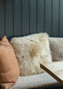 A fluffy cream sheepskin throw pillow centered on a textured beige sofa, flanked by a tan textured pillow on the left and a neutral pillow on the right, set against a dark gray vertical wood-paneled wall with a wooden table edge in the foreground.