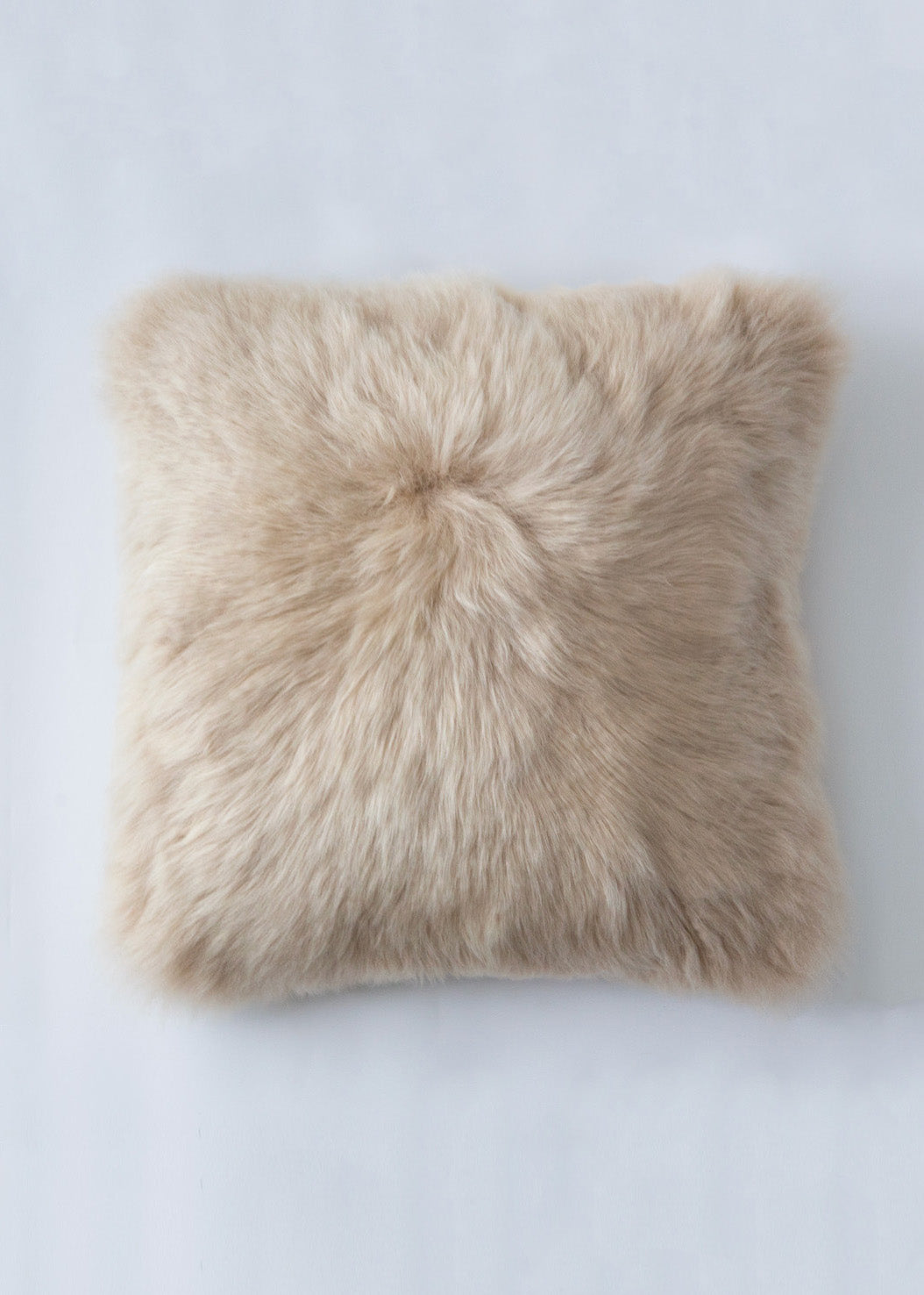 A square sheepskin throw pillow in a light beige color, photographed from directly above on a light gray background.