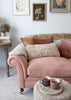 A dusty pink upholstered sofa in a bright, neutral-toned room, styled with sheepskin cream and rust pillows, a sheepskin rug, framed wall art, and a small side table with mugs.
