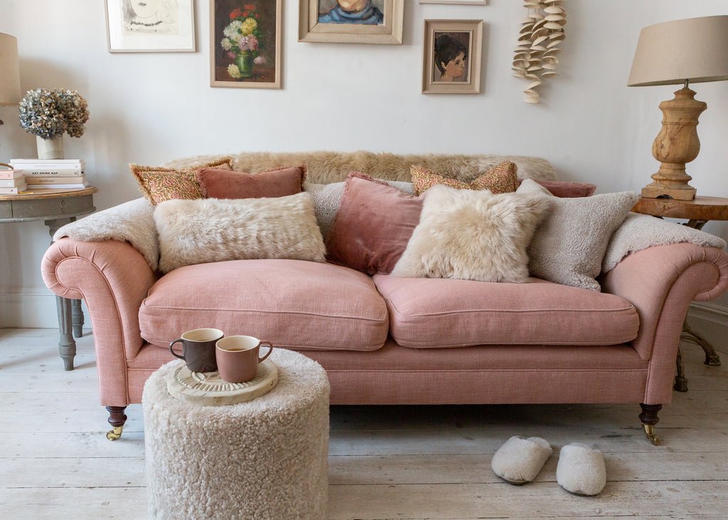 A dusty pink velvet loveseat sofa styled in a cozy, rustic living room with textured cream sheepskin pillows, a matching sheepskin ottoman, and rustic wooden furniture.