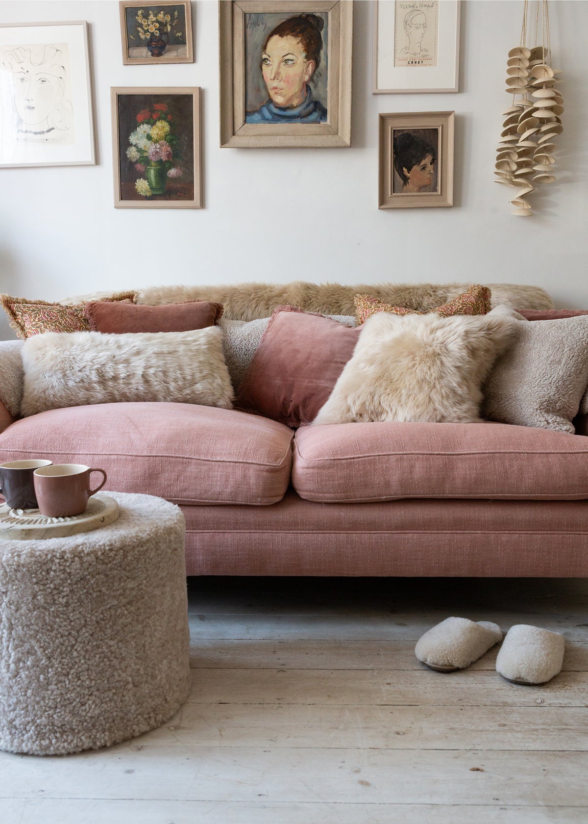 A dusty pink fabric sofa in a cozy living room setting, styled with sheepskin throw pillows, a matching sheepskin ottoman, and neutral slippers on a light wood floor.