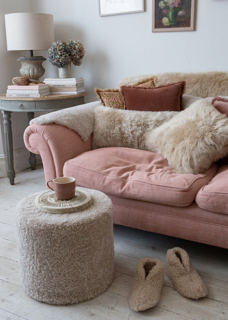 A cozy living room scene featuring a dusty pink velvet sofa adorned with fluffy cream sheepskin and sheepskin pillows, a matching round sheepskin ottoman with a cup on top, and beige slippers on the light wood floor.