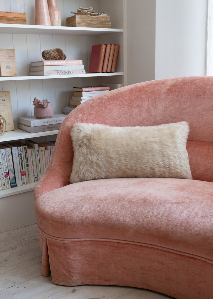 A dusty pink velvet armchair in a bright room, styled with a sheepskin cream throw pillow, with white bookshelves in the background.