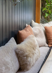 A cozy window seat cushion set with fluffy sheepskin and textured linen pillows in cream, beige, and terracotta, set against a dark gray paneled wall.