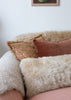 A cozy close-up of a sofa styled with textured throw pillows, including a cream sheepskin pillow, a dusty rose velvet pillow, and a patterned accent pillow on a blush pink couch against a white wall.
