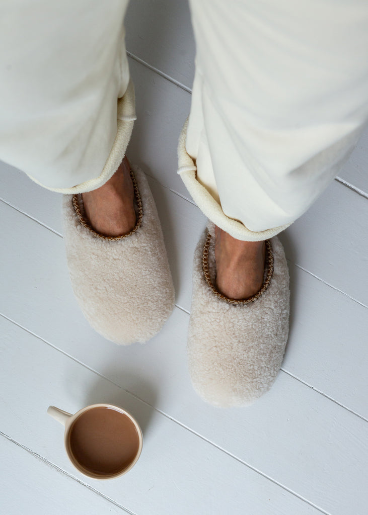 A top-down view of a person wearing fluffy cream-colored slippers with white pants, standing on a light gray wooden floor next to a small ceramic mug.