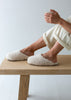 A person sitting on a light wood bench wearing fluffy cream-colored slippers and light pants, holding a white mug against a clean white background.