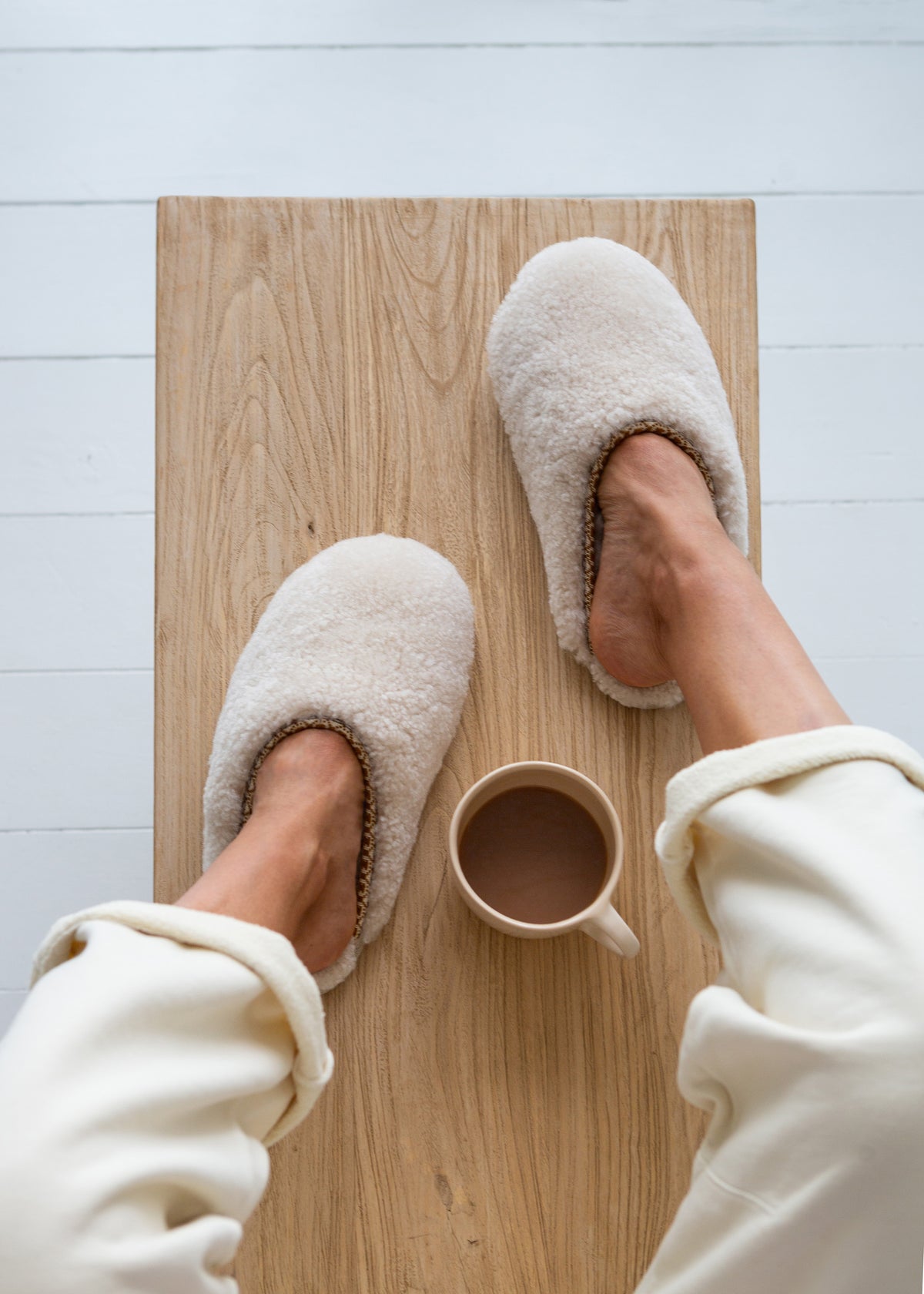 A top-down view of a person wearing cream-coloured sheepskin slippers while sitting on a light wood bench with a small mug of coffee.