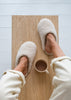 A top-down view of a person wearing cream-coloured sheepskin slippers while sitting on a light wood bench with a small mug of coffee.