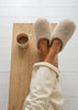A top-down view of a person's legs crossed on a wooden bench, wearing fluffy cream-colored slippers and cream sweatpants, with a matching mug on the bench against a white wood-paneled background.