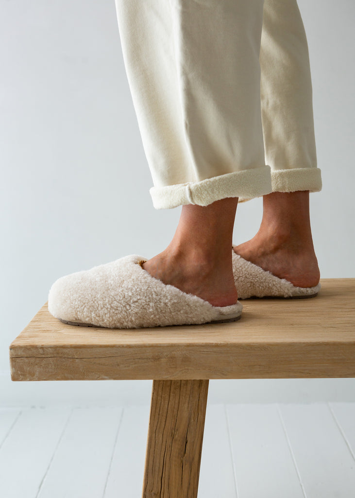 A person wearing cream-colored sheepskin slippers stands on a light wood stool against a clean white background.
