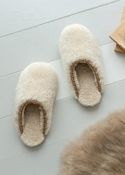 A pair of fluffy cream-colored sheepskin slippers with a braided tan trim, displayed on a light gray wooden floor next to a piece of brown fur.
