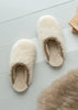 A pair of fluffy cream-colored sheepskin slippers with a braided tan trim, displayed on a light gray wooden floor next to a piece of brown fur.