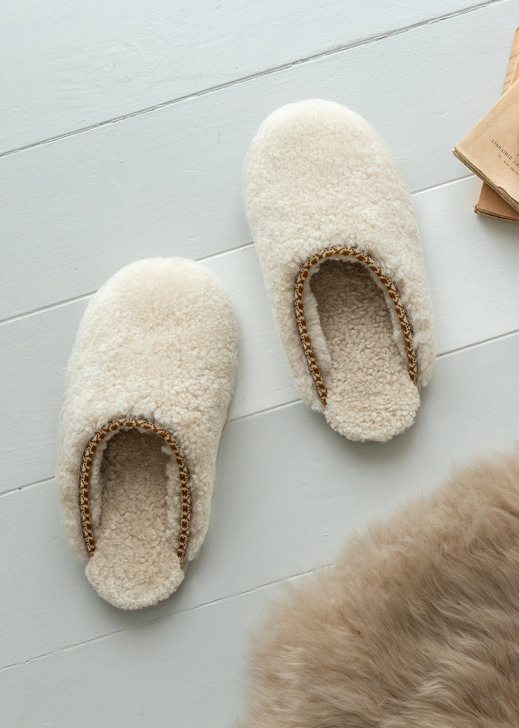A pair of fluffy cream-colored sheepskin slippers with a braided tan trim, displayed on a light gray wooden floor next to a piece of brown fur.