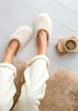 A top-down view of a person wearing fluffy cream-colored slippers with white sweatpants, sitting on a light wood floor next to a cup of coffee and a fluffy beige rug.
