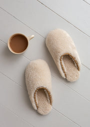 A pair of cream-colored sheepskin slippers with gold trim, displayed on a light gray wooden plank floor next to a ceramic mug filled with coffee.