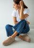 A woman sits on a white floor wearing beige slip-on sheepskin slippers with pink stitching, a white t-shirt, and blue jeans.