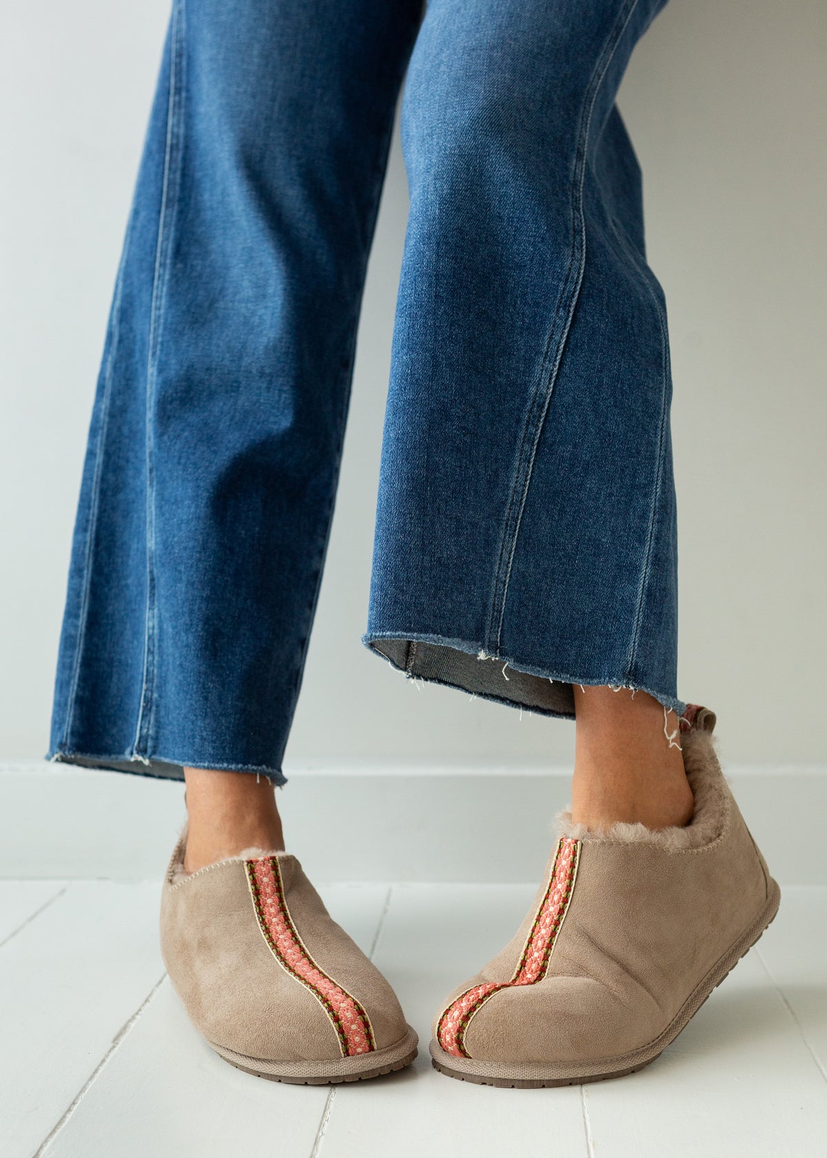 A person wearing blue jeans and beige slip-on sheepskin boots with orange-red decorative trim, standing on a white tiled floor against a white wall.