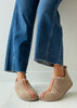 A person wearing blue jeans and beige slip-on sheepskin boots with orange-red decorative trim, standing on a white tiled floor against a white wall.