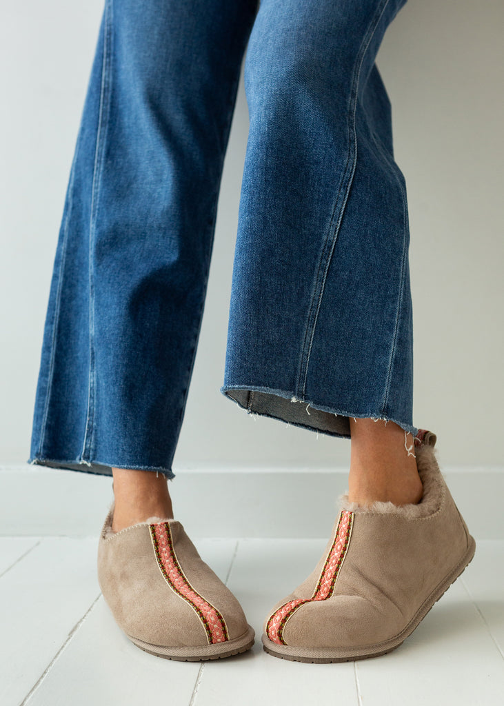 A person wearing blue jeans and beige slip-on sheepskin boots with orange-red decorative trim, standing on a white tiled floor against a white wall.