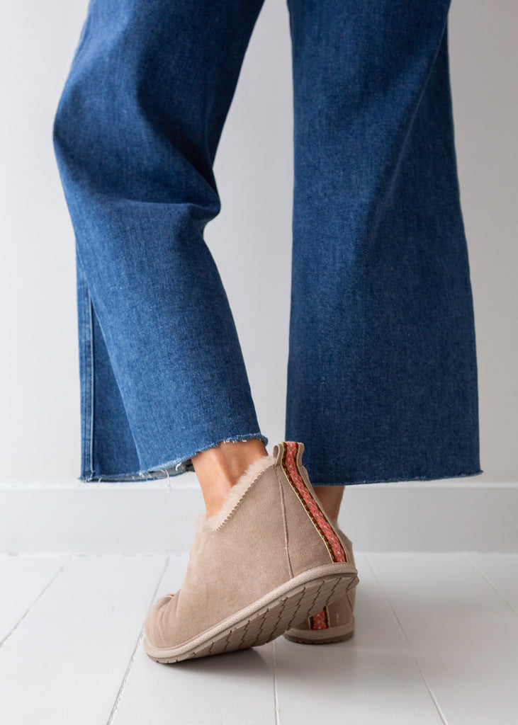 A person wearing blue wide-leg jeans and sheepskin slip-on bootswith a red-orange heel accent, standing on a white floor against a white wall.