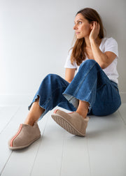 A woman sits on a white wooden floor against a white wall, wearing beige slip-on slippers with red stitching, blue jeans, and a white t-shirt.